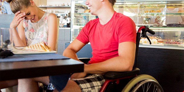 Man i rullstol skrattar med kvinna på ett café. Fotografi